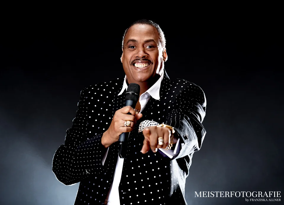 Keith smiling with a microphone in a polka-dot jacket, pointing towards the camera