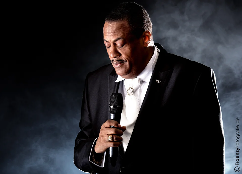 Keith with closed eyes and a microphone, wearing a black suit